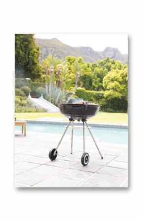 A charcoal grill is ready for a barbecue by a poolside with lush greenery in the background