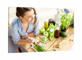 Young and happy woman eating healthy salad sitting on the table with green fresh ingredients indoors