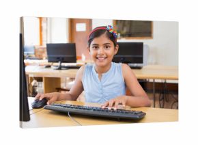 Indian schoolgirl wearing headband smiling at monitor in computer lab using keyboard and mouse