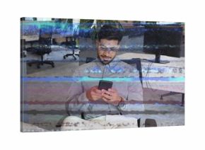 Indian office worker wearing glasses looking at smartphone sitting at desk in office, with monitors