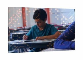 Writing Middle Eastern boy focusing on penning notes at individual school desk, with notebook, pen