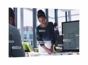 Leaning architect reviewing plans at office table, wearing dark blouse white skirt, with monitor