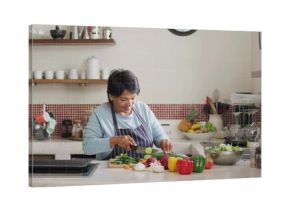 Chopping senior Indian cook in striped apron slicing cucumber at kitchen island, with cutting board