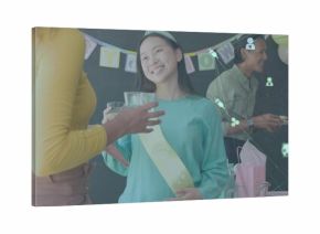 Celebrating woman wearing pale yellow sash and teal top raising clear glass at home with gifts