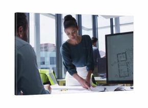 Pointing lead in teal blouse, white skirt leaning over table in office with monitor, vest, plans