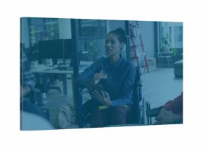 Speaking woman holding tablet and stylus in wheelchair at open-plan office, wearing blue shirt