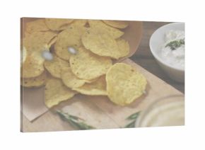 Spilling corn tortilla chips from paper cone onto wooden board on rustic tabletop, with herb dip