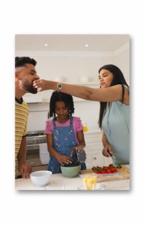 Mother feeding husband while daughter prepares fruit salad in kitchen together