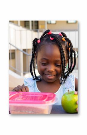 African american schoolgirl at table and having healthy lunch eating apple outside elementary school