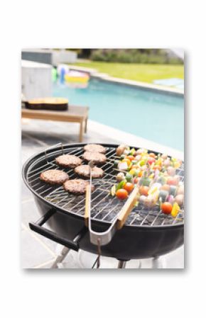 A barbecue grill is loaded with burgers and vegetable skewers, with a pool in the background