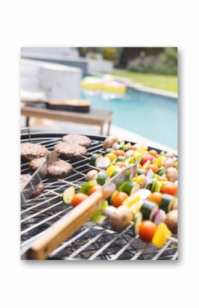 Burgers and vegetable skewers are grilling by a poolside
