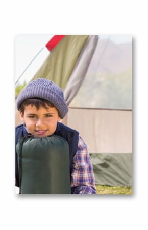 Middle Eastern boy sitting on grass at campsite holding sleeping bag near khaki-red canvas tent