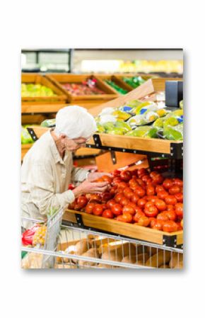 Senior woman examining tomato from bin in produce aisle, pushing cart with avocados and apples