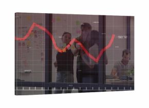 Three coworkers analyzing red line graph in glass-walled meeting area, using sticky notes