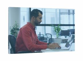 Typing Middle Eastern man in red shirt and dark pants at white desk using silver laptop