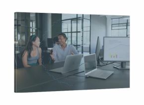 Talking pair wearing sleeveless top and blue shirt at table in office with silver laptops, monitor