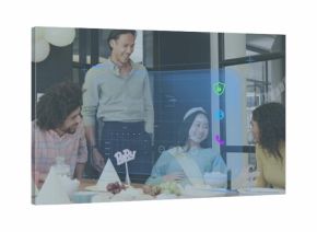 Laughing team leaning toward office table, celebrating guest wearing sash, headband with hologram