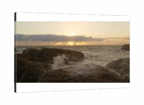 Crashing ocean waves spraying sea spray on jagged shore rocks, with sun rising behind low clouds