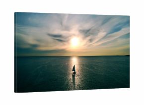 Little white boat floating on the water towards the horizon in the rays of the setting sun. Beautiful clouds with yellow highlights. Aerial view