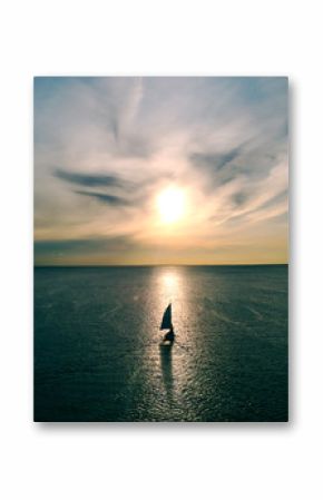 Mała biała łódź unosi się na wodzie w kierunku horyzontu w promieniach zachodzącego słońca. Piękne chmury z żółtymi refleksami. Widok z lotu ptaka