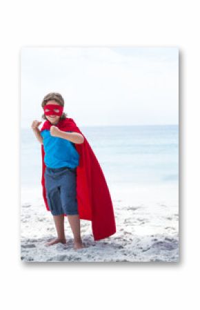 Child wearing red superhero cape and mask punching air on white sandy beach near calm sea