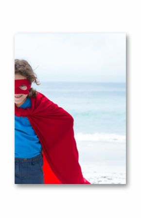 Child is standing on sandy beach wearing bright red cape and eye mask, ocean horizon visible