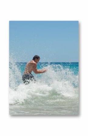 Man in dark swim shorts is wading in crashing turquoise waves at beach, sending water spray