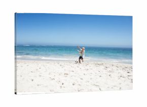 Boy kicking soccer ball at shoreline on white sand near turquoise ocean under clear sky