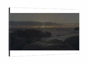 Sun sinking over horizon, casting glow on ocean waves crashing on rocky seashore under cloud layer
