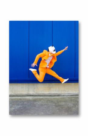 Man wearing vibrant orange suit and panda mask jumping against blue wall