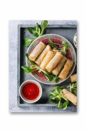 Fried spring rolls with red and white sauces, served in china plate on square wood tray with fresh green salad over gray blue texture background. Flat lay, space. Asian food