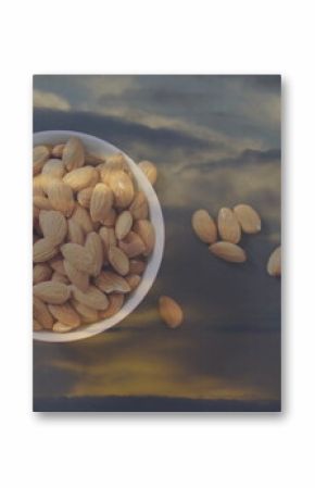 Reflecting white ceramic bowl holding almonds on reflective glass table at dusk, showing cloudy sky