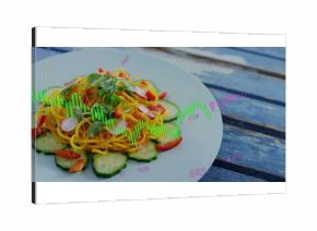 Showing white plate holding yellow spiral noodles, cucumber, pepper and microgreens on blue table