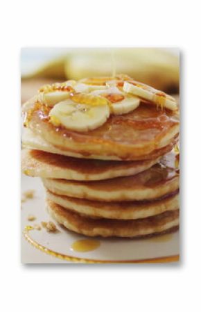 Pancake stack sitting on kitchen table with banana slices and syrup, with granola jar