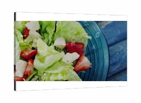 Blue-tinted glass bowl sitting on blue table, holding green lettuce, red strawberries, white cheese