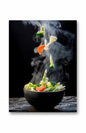 The steam from the vegetables carrot broccoli cauliflower on black bowl , a steaming. Boiled hot Healthy food on table on black background,hot food and healthy meal concept