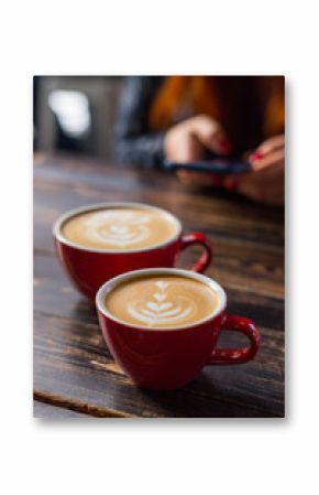 Dwa czerwonej filiżanki kawy na drewnianym tle z latte sztuką. Stolik w kawiarni. Koncepcja łatwego śniadania. Małe i duże ceramiczne kubki. Widok z góry. Dziewczyna z telefonem w tle
