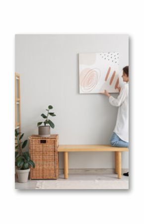Young woman hanging picture on white wall in room