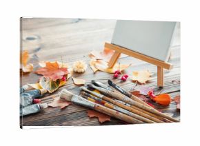 Canvas on easel, paint tubes, brushes for painting and autumn leaves on old wooden background. Retro toned.