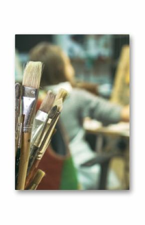 Paintbrushes on the foreground close up and a girl painting a picture on the blurred background. Art lesson in studio.    