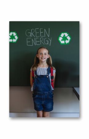 Girl standing in classroom by green board with GREEN ENERGY text, recycle icons, turbine, laptop