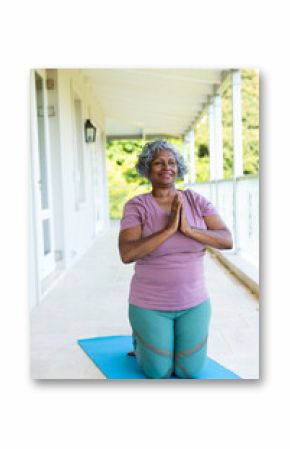 African american senior woman smiling and meditating in prayer pose while kneeling on mat in balcony