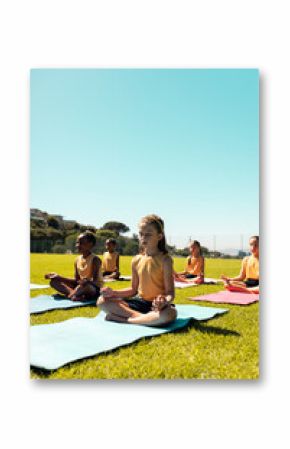 Diverse schoolgirls practicing yoga and meditating in sports field at elementary school, copy space