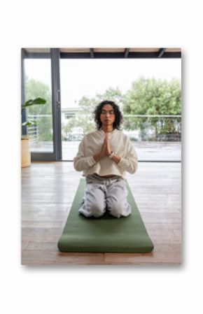 Non-binary person wearing smartwatch kneeling and praying on green yoga mat by sliding glass doors