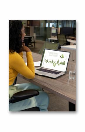 Vertical of biracial businesswoman using laptop in empty office with graph and data on screen