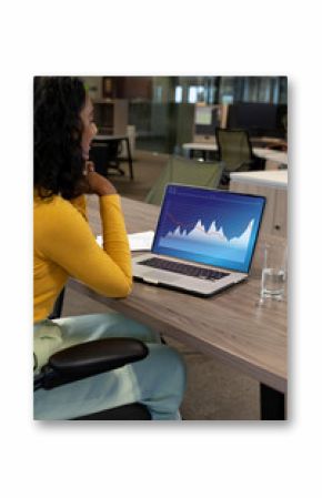 Vertical image of biracial businesswoman using laptop in empty office, with graphs on laptop screen