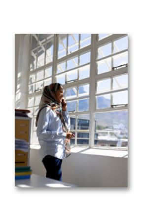 Happy muslim biracial businesswoman in headscarf talking on smartphone at window in modern office