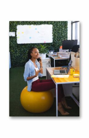 Happy african american casual businesswoman using laptop and talking on smartphone in office