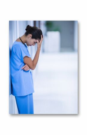 Nurse leaning against wall in bright hospital corridor, holding forehead and wearing stethoscope