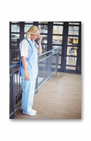 Nurse in scrubs leaning on railing holding forehead with stethoscope in mezzanine near glass doors
